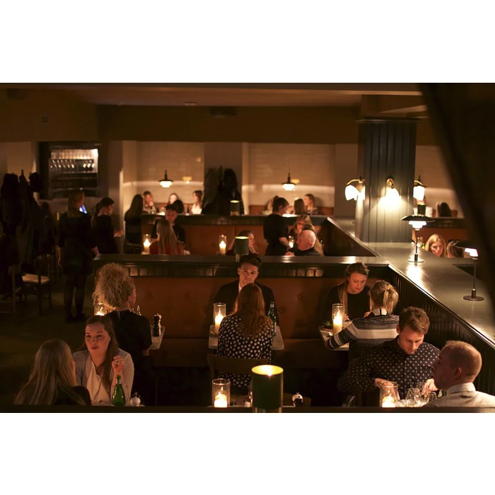 Svagt oplyst restaurant med flere grupper af mennesker, der spiser ved bordene, engageret i samtaler, med stearinlys og flasker på bordene.